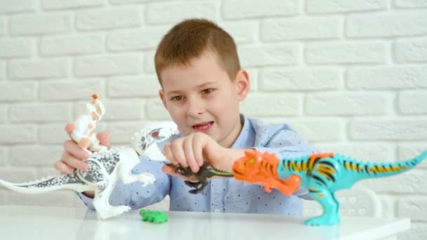 Дитячий хлопчик сидить за білим столом грає в битву з іграшковими динозаврами . — стокове відео