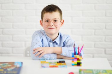 Beyaz bir masada oturan gözlüklü bir öğrenci defter ve kitapların arka planına gülümsüyor..