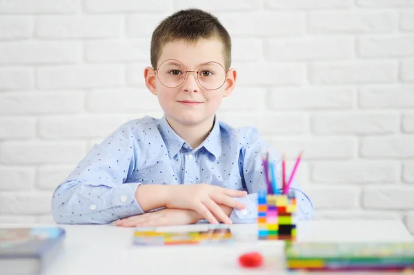 Anak sekolah dengan kacamata duduk di meja putih tersenyum terhadap latar belakang notebook dan buku. — Stok Foto