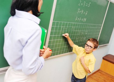 Gözlüklü küçük bir çocuk tahtaya tebeşirle yazı yazar ve sınıfın arkasındaki öğretmene bakar..