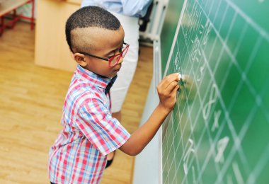 Küçük bir çocuk. Kırmızı gözlüklü bir Afro-Amerikan öğrenci. Yeşil bir karatahtaya tebeşirle yazı yazıyor..