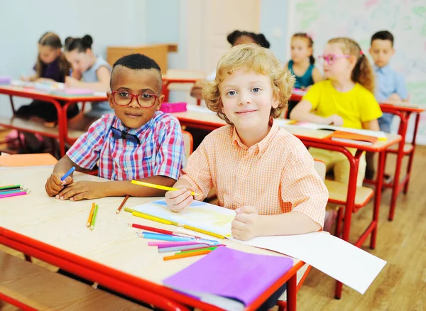 Dos niños pequeños de estudiantes de primaria se están preparando para una lección sentados en un escritorio con lápices de colores en sus manos — Foto de Stock