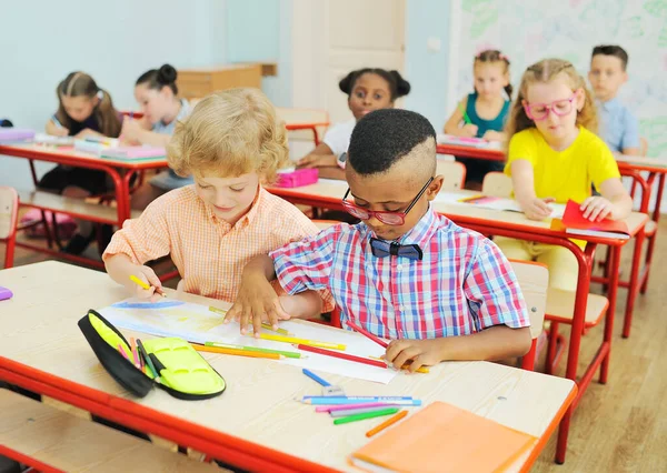 Dos niños pequeños de estudiantes de primaria se están preparando para una lección sentados en un escritorio con lápices de colores en sus manos — Foto de Stock