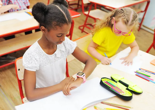 Seorang gadis Afrika-Amerika kecil duduk di sebuah meja di sekolah dasar melihat jam tangan pintar di tangannya dengan latar belakang kelas sekolah dan anak-anak. — Stok Foto