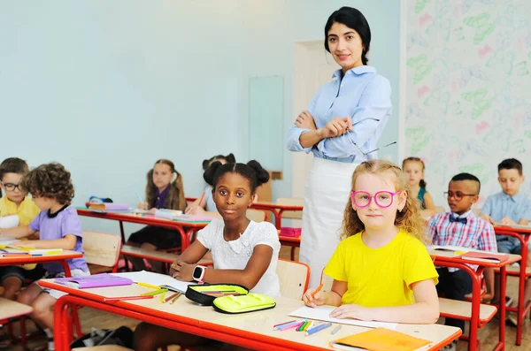 Dos niños-una niña afroamericana y una niña caucásica en gafas de color rosa sonríen sentados en un escritorio en una escuela primaria en el contexto de un maestro y un grupo de niños de la escuela en el — Foto de Stock