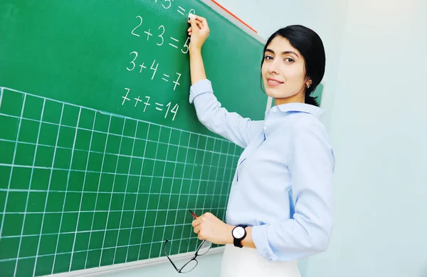 Jovem professora atraente escreve exemplos matemáticos com giz em um quadro-negro verde. De volta à escola, dia dos professores, dia do conhecimento, aritmética, escola primária. — Fotografia de Stock