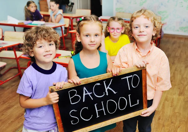 Kelompok anak-anak sekolah tersenyum sambil memegang papan bertuliskan "Kembali ke sekolah" dengan latar belakang ruang kelas dan anak-anak yang duduk di meja sekolah dasar atau sekolah dasar. — Stok Foto
