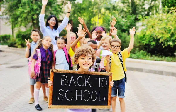 Seorang anak laki-laki kecil berambut keriting tersenyum dan memegang papan dengan tulisan "Kembali ke sekolah" dengan latar belakang sekelompok anak-anak dari kelas yang sama dan seorang guru perempuan. — Stok Foto