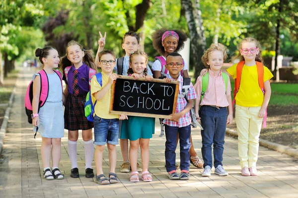 Kelompok anak-anak sekolah multi-ras dengan pakaian berwarna-warni, membawa tas sekolah dan ransel memegang tanda yang bertuliskan "Kembali ke sekolah" dengan latar belakang hijau dan taman. — Stok Foto