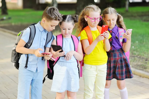 Sekelompok anak-anak sekolah dengan pakaian berwarna-warni dengan tas sekolah dengan smartphone di tangan mereka pergi ke sekolah. Kembali ke sekolah gadget, hari pengetahuan, ketergantungan pada ponsel. — Stok Foto