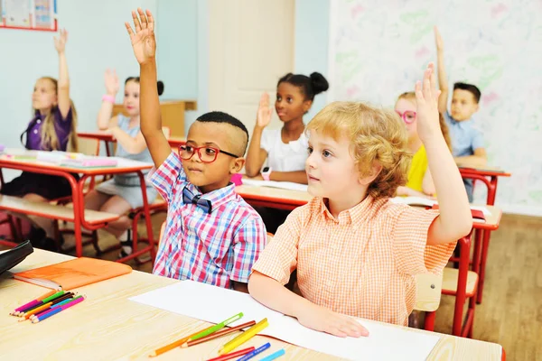 Dos niños pequeños de la escuela levantan la mano para responder a una tarea de maestros en una escuela junior en el contexto de un aula — Foto de Stock