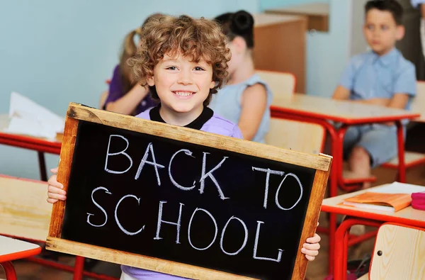 Seorang anak kecil seorang anak sekolah dengan rambut keriting tersenyum terhadap latar belakang sekelompok anak-anak dan kelas sekolah memegang tanda yang bertuliskan "Kembali ke sekolah". Hari pengetahuan, 1 September, pendidikan — Stok Foto