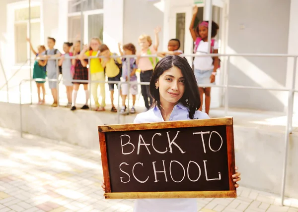 Söt kvinnlig lärare med en skylt som säger "tillbaka till skolan" på bakgrunden av barn och grundskolan. — Stockfoto