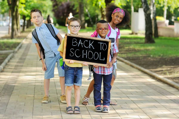 Kelompok anak-anak sekolah multi-ras dengan pakaian berwarna-warni, membawa tas sekolah dan ransel memegang tanda yang bertuliskan "Kembali ke sekolah" dengan latar belakang hijau dan taman. — Stok Foto