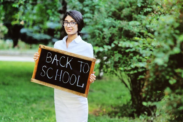 Joven profesora en ropa elegante y estricta gafas con un cartel "Volver a la escuela" sonríe en el fondo de la vegetación en el Parque. El día del maestro, el día del conocimiento, la educación. — Foto de Stock