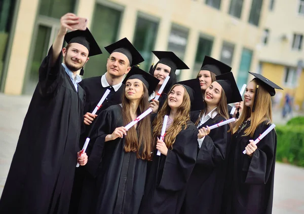 group of graduate students of masters in black robes and square hats take selfies on a smartphone camera.