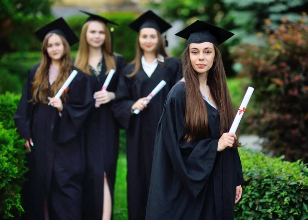 Jong meisje, een afgestudeerde student in een zwart gewaad en een vierkante hoed met een diploma in haar handen, glimlacht tegen de achtergrond van studenten klasgenoten. — Stockfoto