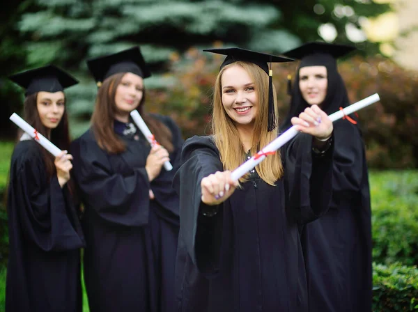 Bonita joven es una estudiante universitaria graduada en una túnica negra sonriendo sosteniendo un diploma en sus manos en el fondo de un grupo de graduados. —  Fotos de Stock