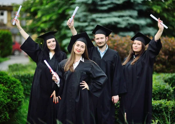 Bonita joven es una estudiante universitaria graduada en una túnica negra sonriendo sosteniendo un diploma en sus manos en el fondo de un grupo de graduados. —  Fotos de Stock