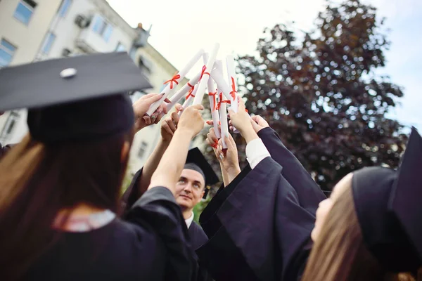 Grupo de graduados universitários em vestes de estudante ou manto e chapéus quadrados na cerimônia de formatura realizar certificados de graduação da faculdade em suas mãos e se alegrar. — Fotografia de Stock