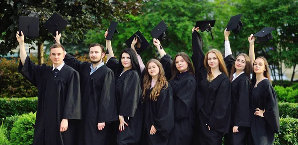 Csoport egyetemi vagy főiskolai diplomások fekete köpenyben felemelte a kezét négyszögletes sapkák diplomások és örülnek a diploma. — Stock Fotó