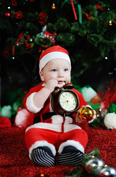 Child dressed as Santa Claus gnawing teeth clock alarm clock. — Stock Photo, Image