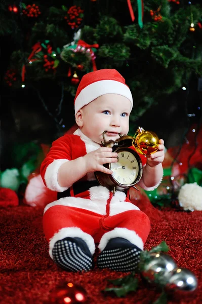 Child dressed as Santa Claus gnawing teeth clock alarm clock. — Stock Photo, Image