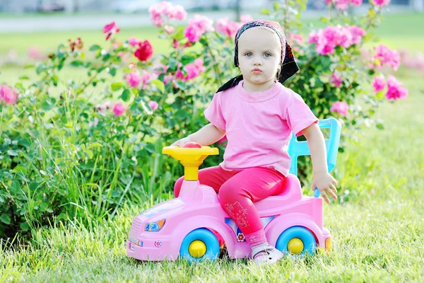 Babymädchen fährt rosa Spielzeugauto auf einem Hintergrund aus Rosen — Stockfoto