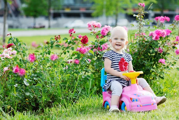 Babymädchen fährt rosa Spielzeugauto auf einem Hintergrund aus Rosen — Stockfoto