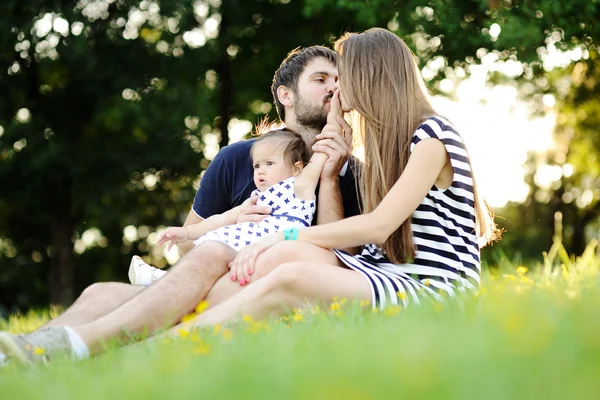 Молодая семья отдыхает в парке на траве. Мама и папа целуются — стоковое фото