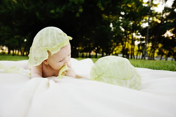 Baby girl with cabbage. Cabbage leaves on the head of a child. Child relaxing in a park on the grass. — Stock Photo, Image