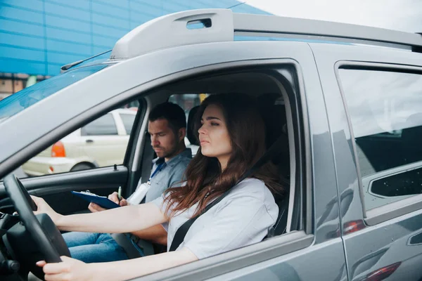 Giovane donna sconvolta prende esame di guida con grave istruttore di auto maschile. Due persone sedute in auto grigia con cinture di sicurezza allacciate. Concentrato giovane donna tiene volante e guarda avanti. — Foto Stock