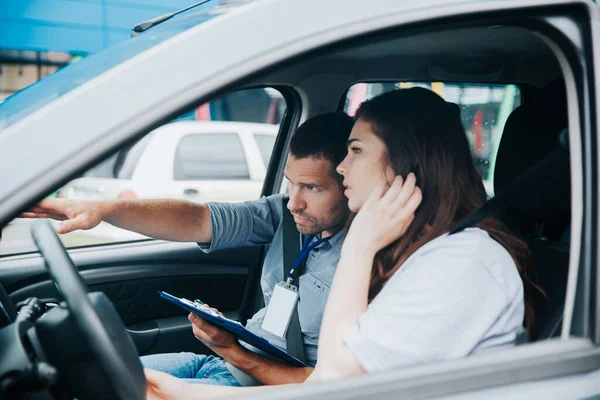 Maschio istruttore di auto spiega le regole di guida per la studentessa. L'uomo caucasico indica la strada e tiene il suo giornale. Giovane donna fissa i capelli e ascolta i commenti sui suoi errori. — Foto Stock