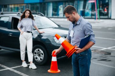 Erkek yorgun sürücü eğitmeni mesai bitiminde trafik konileri topluyor. Arka planda siyah arabanın yanında duran hafif günlük kıyafetli, bulanık kadın. Yağmurdan sonra ıslak asfalt.