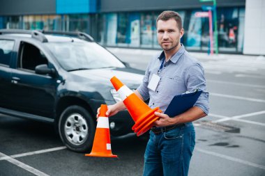 Sürücü eğitmeni işini seviyor. Genç ve çekici bir adam elinde pano ve koni ile kameraya bakıyor. Arka planda siyah araba var. Sürücü ehliyeti almak.