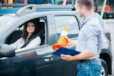 Kot pantolon ve gri gömlekli erkek eğitmen bayan öğrencisine yol konisi gösteriyor. Çekici bayan öğrenci onun açıklamalarını dinlerken gülümsüyor ve arabanın camından bakıyor. Sürücü okulu konsepti.
