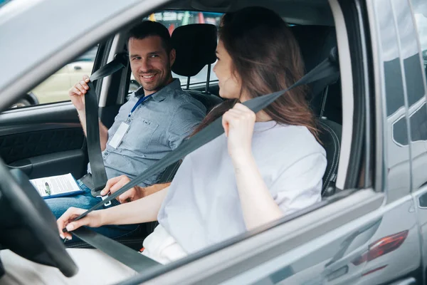 Giovane studentessa e attraente istruttore di guida maschile si sorridono a vicenda seduti in macchina e allacciando le cinture di sicurezza. Esperienza positiva con il concetto di corsi di guida. — Foto Stock