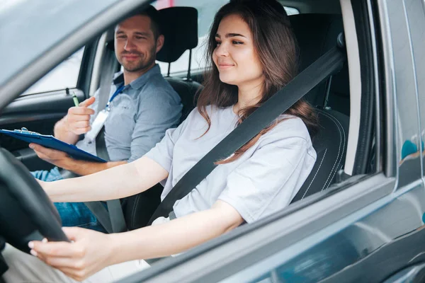 Instrutor do sexo masculino satisfeito com o passeio de sua aluna, ele sorri e comenta sobre suas ações corretas. Duas pessoas estão andando em um carro cinza e usando cintos de segurança. — Fotografia de Stock