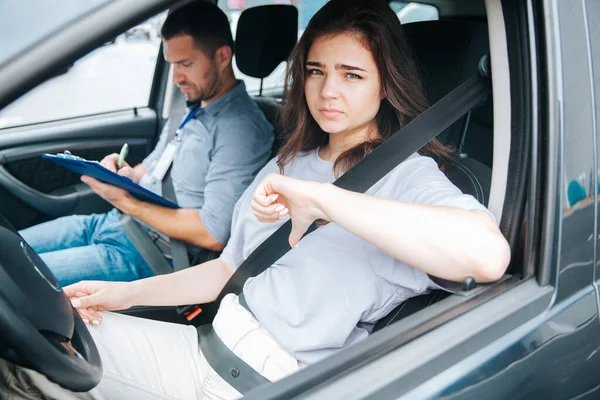 Estudante triste falha em seu teste de condução e mostra os polegares para baixo. Mulher atraente senta-se ao volante de um carro e olha para fora da janela lateral. Instrutor do sexo masculino no banco do passageiro da frente toma notas. — Fotografia de Stock