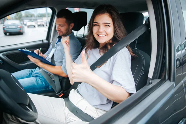 Giovane donna felice pollice in su. Studentessa sorride a causa di un test di guida di successo, guarda la fotocamera e punta su istruttore di guida maschile per mano. — Foto Stock