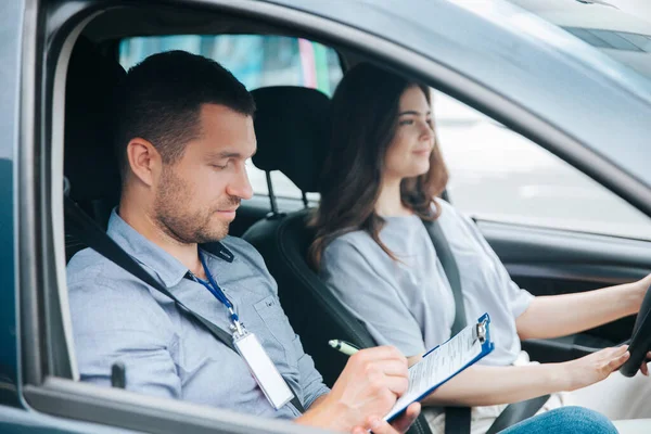 Imparare a guidare un'auto. Istruttore e studentessa di sesso maschile che praticano la guida. Il giovane nota i risultati del test nel suo articolo. Donna allegra soddisfatta delle lezioni di guida. — Foto Stock