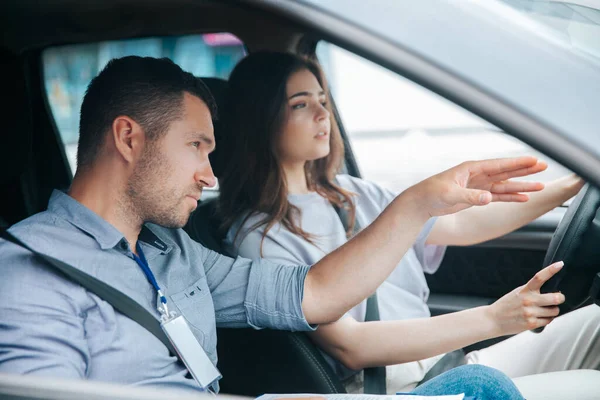 Male instructor showing something by his hand on the road and giving advice. Attractive woman listening her teacher, looking ahead with concentration and holding wheel. — Stock Photo, Image