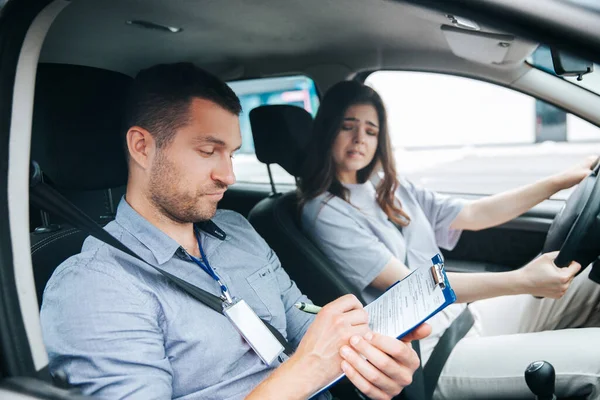 Upset woman failed driving exam bites lips and worries about it. Male instructor notice results of test in his paper. Female student holding wheel and looking troubled at the driving instructor.