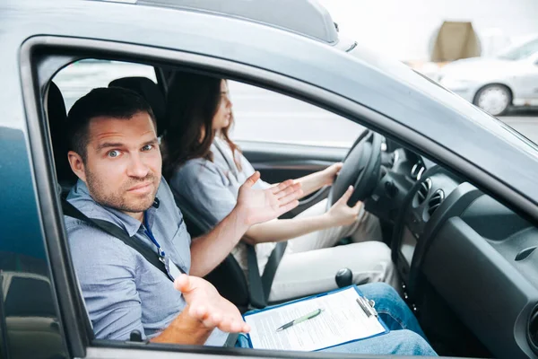 Male driving instructor shrugs up and looks at camera. Man doesnt understand what his female student is doing. Attractive blurred woman sits on driving seat, holds wheel and tries to concentrate on