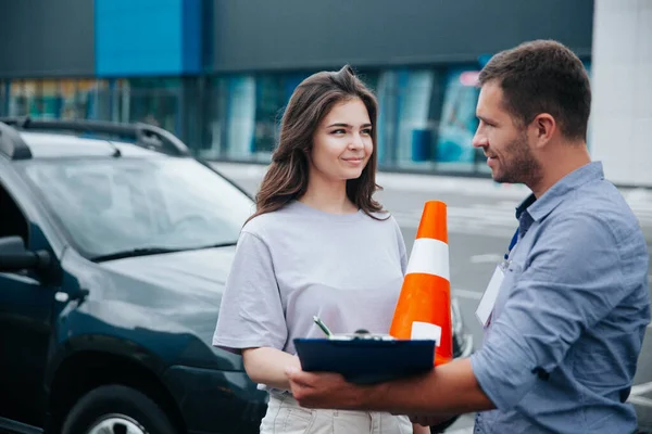 Vrouwelijke student van de rijschool tekent instructeurs papieren en kijkt naar hem met belangstelling. Oranje verkeerskegels met witte streep. Rijlessen. Het verkrijgen van een rijbewijs concept. — Stockfoto