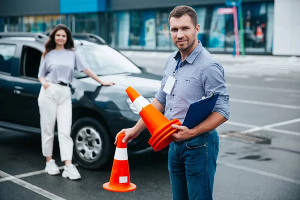 Boldog férfi vezető oktató kezében közlekedési bójákkal, kamerába néz és mosolyog. Fiatal nő áll az autó mellett, és vár, amikor a férfi befejezi a munkáját.. — Stock Fotó