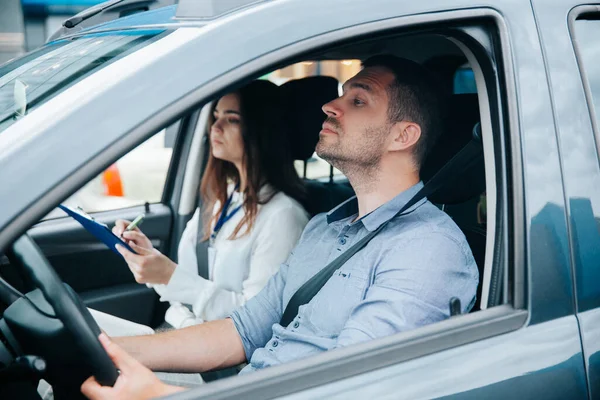 Giovane prende esame di guida con grave istruttore di guida femminile. Studente maschio concentrato tiene volante e guarda avanti. Due persone sedute in auto grigia con cinture di sicurezza allacciate. — Foto Stock