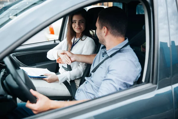 Istruttore di auto femminile che stringe la mano agli studenti maschi. Donna attraente si congratula con successo esame di patente di guida. Uomo in camicia blu tenendo la ruota per mano e guardando la donna. — Foto Stock