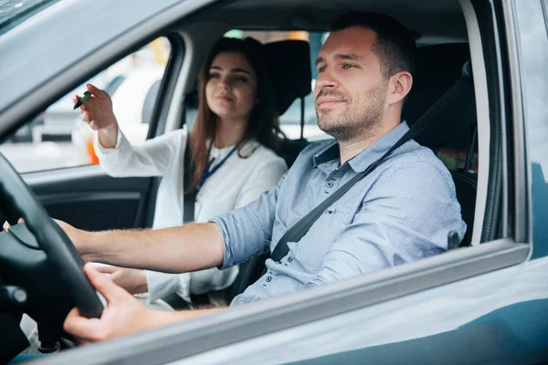 Female driving instructor pointing at object on road by her hand. Diligent male student driving the car and listening what his instructor is saying. Two people in car. Driving lessons concept.