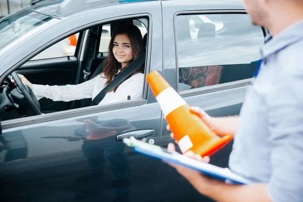 Positive experience in driving school. Cheerful confident young woman is glad for improvement of her driving skills. Male instructor stands near the car holding orange road cone and clipboard.